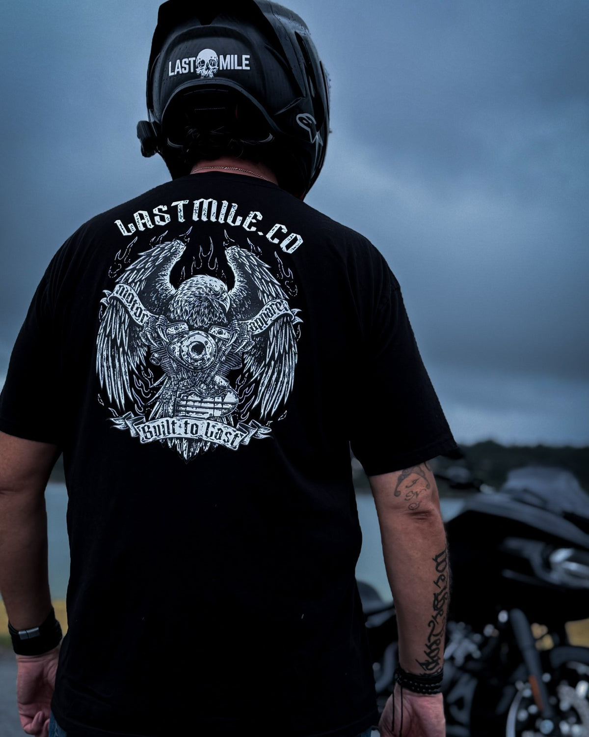 Wing and Iron Tee, worn by a rider, back graphic under storm clouds beside a touring bike