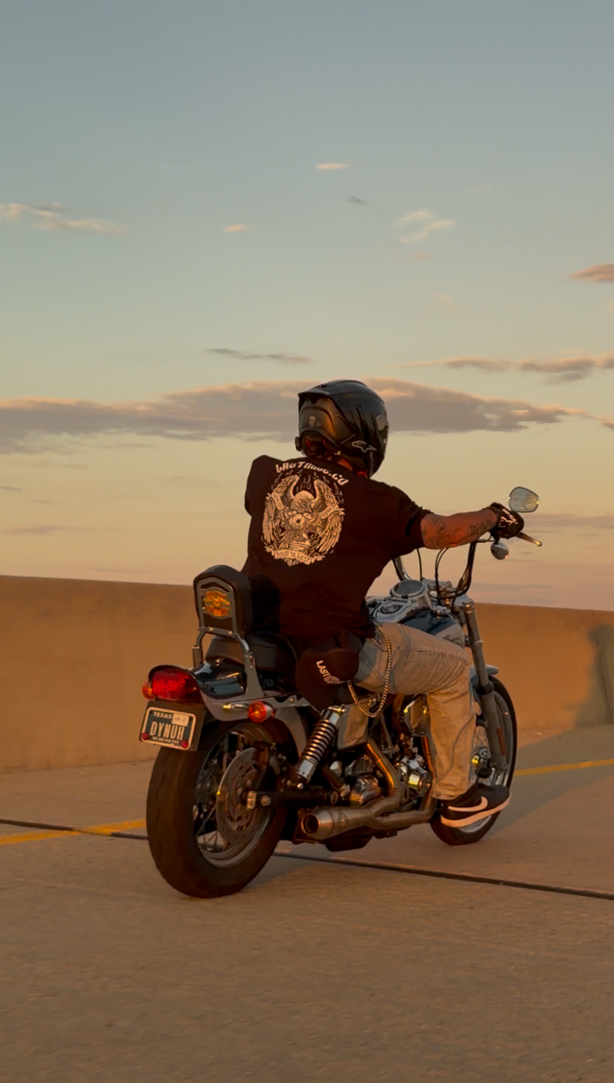 Rider on a Dyna at golden hour wearing the Wing and Iron Tee, eagle and V-twin back print across the shoulders